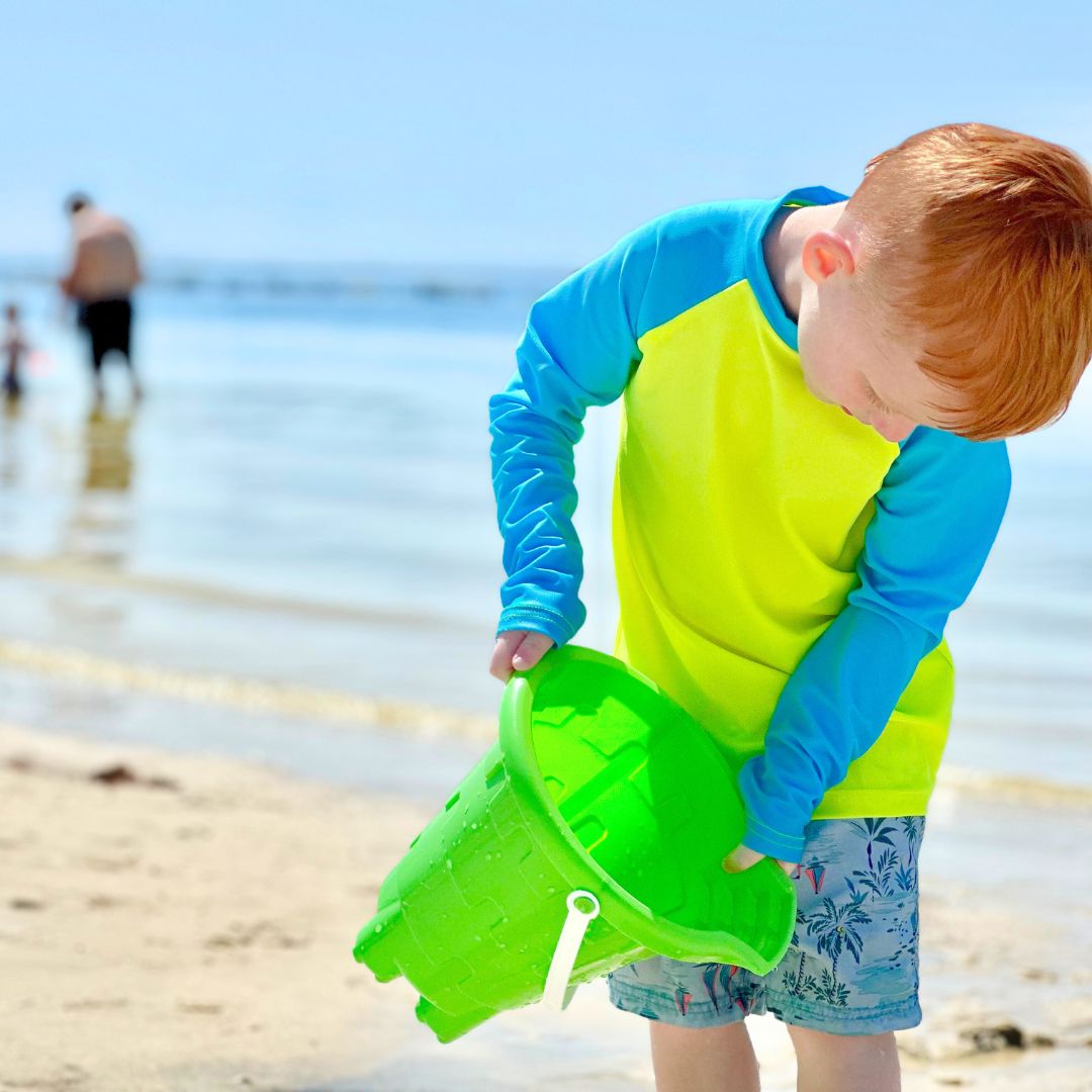 Red haired pre-school boy with bright prneon rash guard sun protective shirt pouring water from a green bucket at the beach.